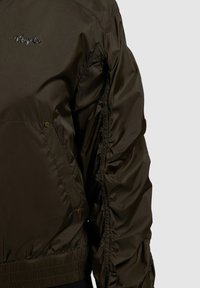 Veste en nylon marron foncé avec poignets élastiques, poche latérale à bouton-pression et logo brodé "Khujo Co" sur la poitrine.