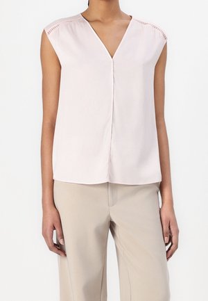 Woman wearing a sleeveless light pink V-neck blouse with delicate shoulder cutout details and beige wide-leg pants.