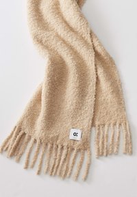 Sciarpa lavorata beige con una finitura morbida e strutturata, caratterizzata da bordi frangiati e un'etichetta bianca con scritte nere su un'estremità.