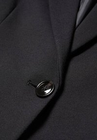 Tissu de blazer noir avec un gros plan sur un bouton noir à quatre trous et la couture de la boutonnière autour.