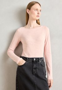 Žena sa ravnom plavom kosom, koja nosi zlatne okrugle naušnice, svijetloružičastu majicu dugih rukava i crnu trapericu viskog struka, s jednom rukom u džepu.