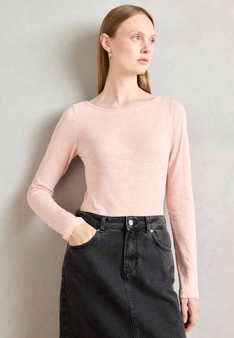 Žena sa ravnom plavom kosom, koja nosi zlatne okrugle naušnice, svijetloružičastu majicu dugih rukava i crnu trapericu viskog struka, s jednom rukom u džepu.