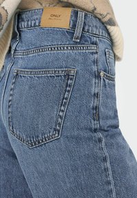 Vue de dos d'un jean en denim bleu avec une étiquette en cuir portant l'inscription "ONLY Blue Denim", porté avec un pull beige.