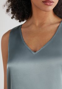 Sáténový top s V výstřihem v jemně šedé barvě, který se vyznačuje hladkým povrchem a dokonalým zakončením kolem výstřihu. Detailní záběr zaměřený na látku.