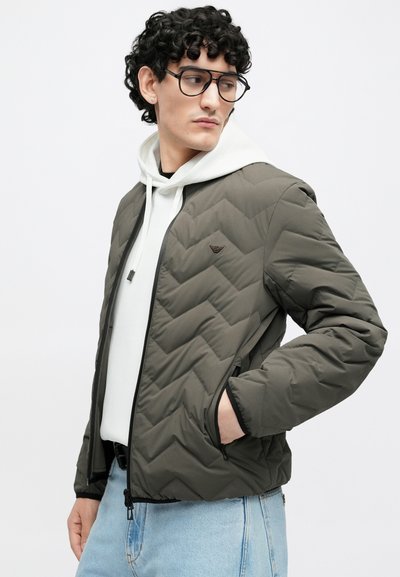 Sivá prešívaná bunda s textúrovaným vzorom šikmých prúžkov, čiernym lemovaním, prednými vreckami a zapínaním na zips, vrstvená cez bielu mikinu s kapucňou.