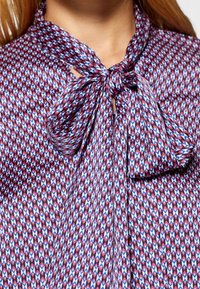 Kortärmad blus i mönstrat tyg med röda, blå och vita geometriska former, med knytning i halsen och slät textur.