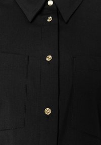 Schwarze Hemdjacke aus einem Stoff mit glatter Textur. Verfügt über goldene Metallknöpfe und zwei Vordertaschen. Klassisches Kragendesign.