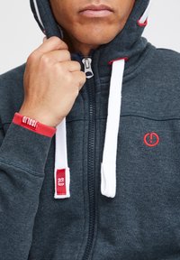 Sweat à capuche zippé gris foncé avec cordons blancs, accents rouges et détail logo. Tissu lisse et doux avec poignets côtelés et fermeture éclair.