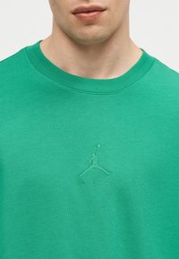 Κοντινό ενός ατόμου που φοράει πράσινο μπλουζάκι με στρογγυλή λαιμόκοψη, με κεντημένο το λογότυπο Jumpman και τη λέξη «AIR» από κάτω στο στήθος.