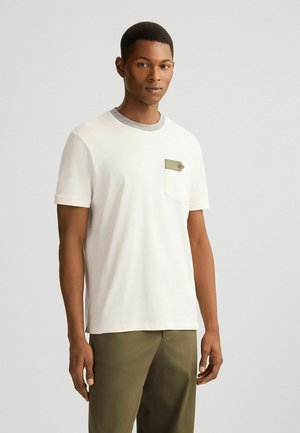 Kurze weiße Baumwoll-T-Shirt mit grauem Halsausschnitt und einer grünen aufgesetzten Tasche mit Knopfdetail auf der linken Brust.
