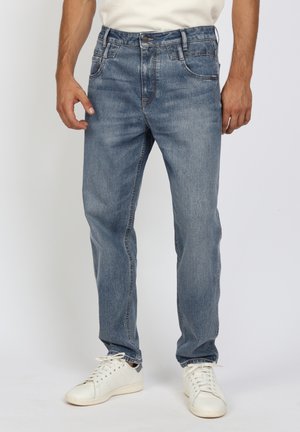 Man wearing blue straight-fit jeans and white sneakers standing against a plain background, hands relaxed by sides.