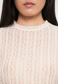 Top di pizzo crema con un motivo a onde testurizzato, caratterizzato da un collo arricciato e tessuto trasparente. Il design è leggero e decorativo.