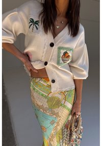 Femme portant un cardigan blanc orné de patchs de fruits et de palmiers, une jupe à motifs colorés, et tenant un sac tressé avec des perles.