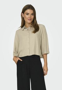Beige, kortærmet skjorte med knapper og krave, lavet af et letvægts, struktureret stof og med løstsiddende ærmer, parret med sorte bukser.