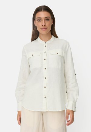 Frau mit langen braunen Haaren, die ein weißes Hemd mit Brusttaschen und hellbeige Hosen trägt, steht vor einem schlichten Hintergrund.