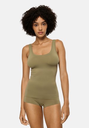 Kvinde med krøllet hår iført en tætsiddende olivengrøn tanktop og matchende boyshorts, stående mod en ensfarvet hvid baggrund.