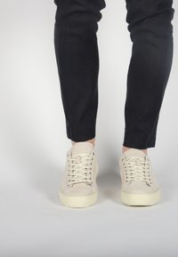 Beige suede sneakers with flat laces and rounded toe. Thick white rubber sole and minimal stitching detail. Worn with dark slim jeans.