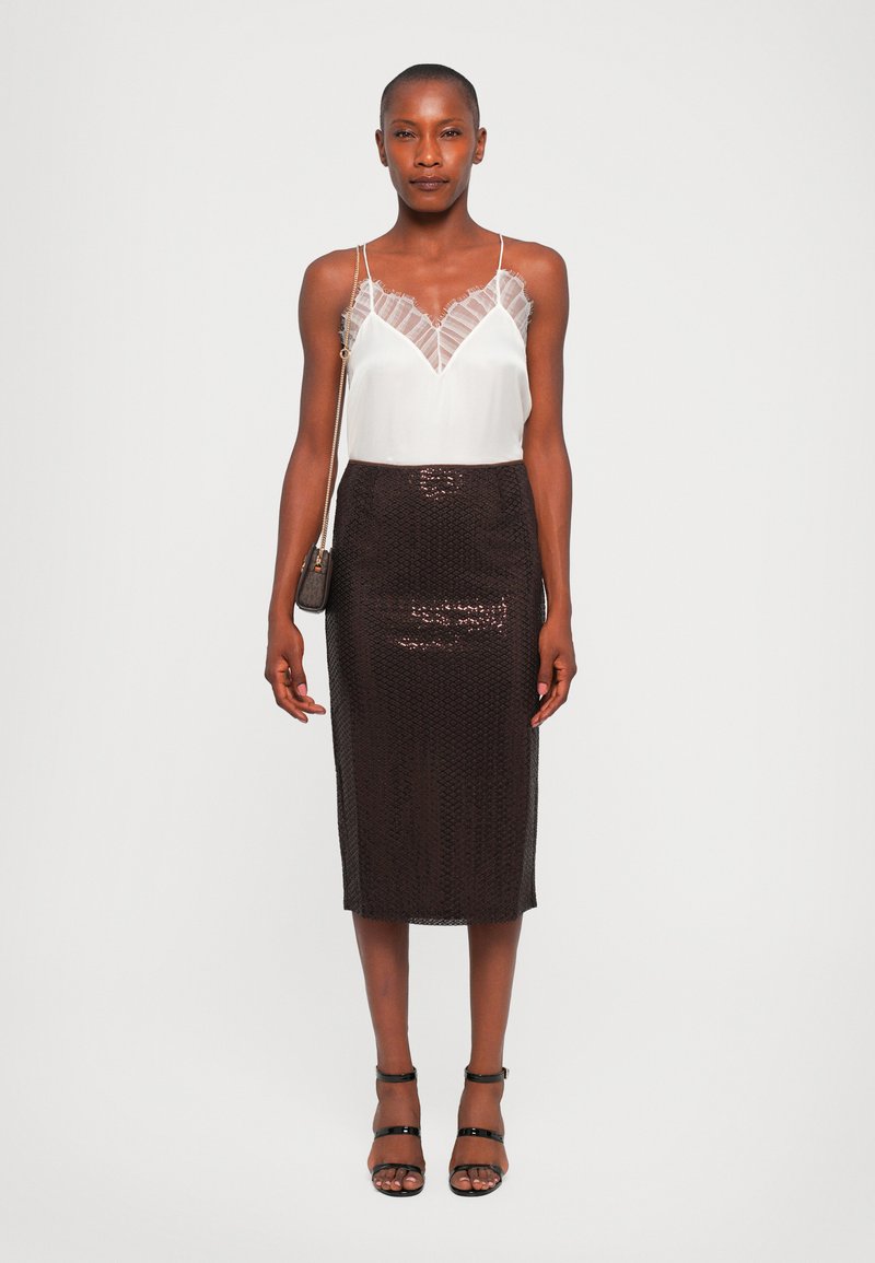 Witte cami met kanten afwerking, gecombineerd met een fitted, donker gekleurde paillettenkokerrok. Model draagt strappy zwarte hakken en houdt een kleine tas vast.