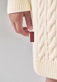 Kreminės spalvos kabeline mezgimo megztinis su ribotomis rankogalių apykaklėmis, ant krašto pritvirtinta raudona etiketė „SUPERDRY“. Ranka švelniai liečia drabužį.