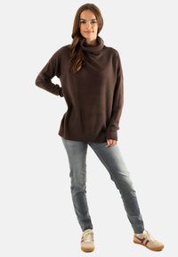 Maglione a collo alto marrone con bordi a costine, vestibilità rilassata, abbinato a jeans azzurri chiari e sneakers beige con dettagli rossi.