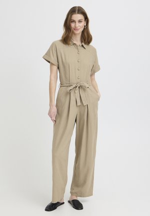 Naine seisab, kandes beeži värvi lühikeste varrukatega nööpidega kombineeritud pükse, vööga seotud vöökohaga ja mustade libisemisvastaste jalanõudega, tavalise tausta ees.