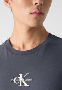 Pilkas medvilnės marškinėliai su apvalia iškirpte ir baltu išsiuvinėtu logotipu, kuriame parašyta "Calvin Klein Jeans", ant krūtinės. Trumpomis rankovėmis.