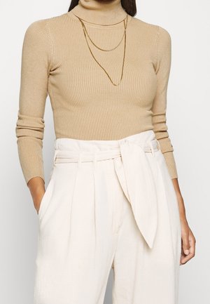 Femme portant un pull à col roulé côtelé beige, des colliers dorés superposés et un pantalon blanc taille haute avec une ceinture en tissu nouée.