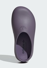 Zapatilla morada de Adidas tipo slip-on con superficie lisa, forma redondeada y diseño recortado. Presenta un logotipo grabado en la parte superior.