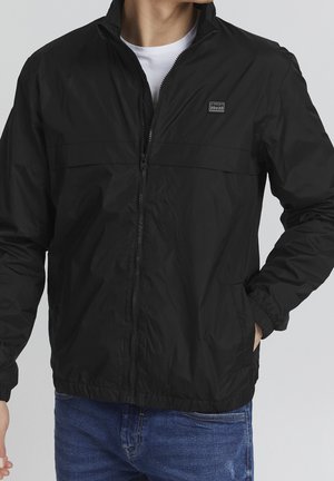 Homme portant une veste noire zippée avec un petit logo sur la poitrine et un jean bleu, une main dans la poche, t-shirt blanc visible en dessous.