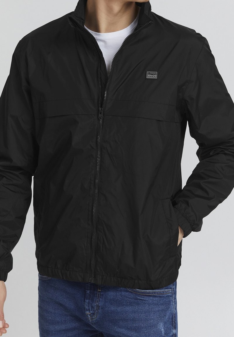 Homme portant une veste noire zippée avec un petit logo sur la poitrine et un jean bleu, une main dans la poche, t-shirt blanc visible en dessous.