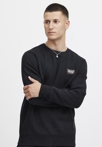 Svart sweatshirt med rund hals, ribbade muddar och nederkant, med en liten rektangulär logopatch på bröstet. Slät textur.