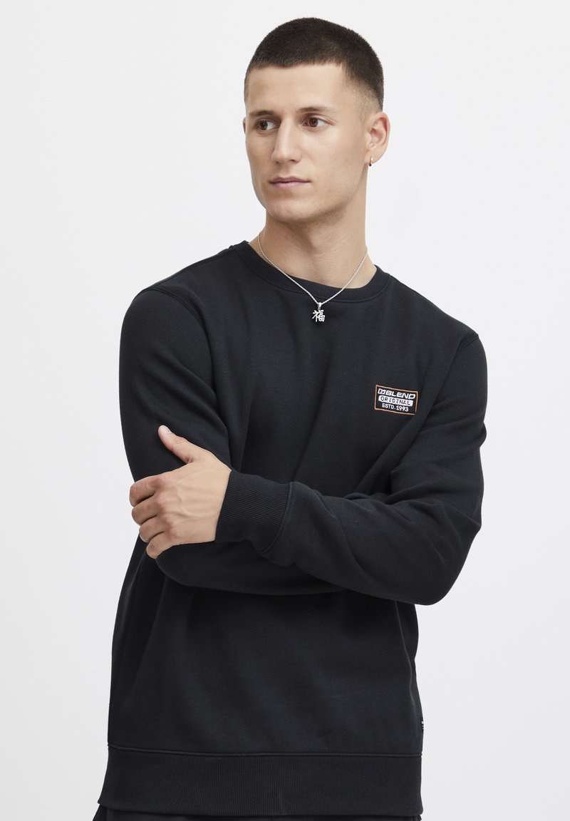 Svart sweatshirt med rund hals, ribbade muddar och nederkant, med en liten rektangulär logopatch på bröstet. Slät textur.