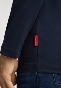 Marineblaue Langarmbluse aus weichem Baumwollstoff, mit gerundetem Saum und einem roten Joop Jeans-Tag an der Seitennaht.