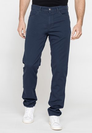 Uomo con pantaloni casual blu navy e sneakers bianche, in piedi davanti a uno sfondo bianco semplice.