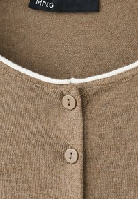 Gros plan sur un cardigan en maille beige avec deux boutons assortis et une bordure blanche le long de l'encolure.