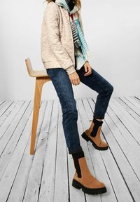 Světle hnědé kotníkové boty s černými látkovými bočními panely, nošené s tmavými džíny a světle prošívanou růžovou bomber bundou, doplněné vzorovaným šátkem.