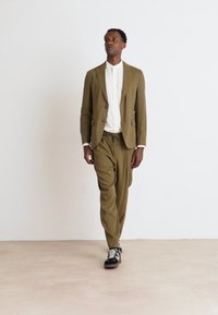Olive green suit with a tailored blazer and relaxed trousers, paired with a white shirt and black sneakers. Minimalist design and smooth texture.