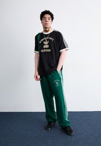 Črna majica Adidas z belimi akcenti in logotipom; zelene hlače Adidas za tek z belimi črtami in detajlom besedila. Črne čevlje.