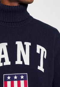 Marineblauer Strickrollkragenpullover mit auffälligen weißen Buchstaben und einem mehrfarbigen Emblem mit Sternen und Streifen auf der Brust.