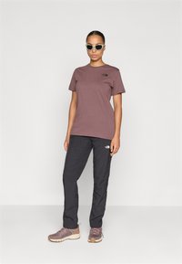 Bordeauxrood t-shirt met korte mouwen en logo, zwarte lichte broek en bruine sportschoenen. Model draagt witte zonnebril. Effen achtergrond.