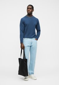 Homme portant un pull bleu à col rayé, jean bleu clair, baskets blanches, tenant un sac fourre-tout noir, debout devant un fond uni.