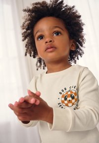 Hellbeiges Langarmhemd mit einer Grafik von einem Kürbis und einem Schachbrettmuster auf der Vorderseite, gefertigt aus weichem Stoff mit rundem Ausschnitt.