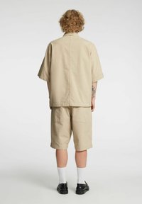 Beige kortermet skjorte og shorts sett med teksturert mønster, avslappet passform og lappelommer. Brukes med hvite sokker og svarte sko.