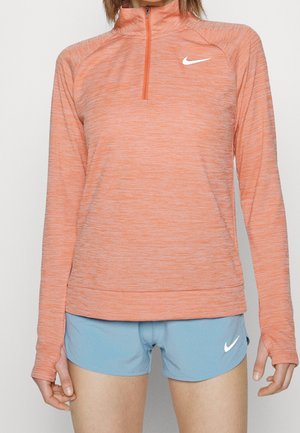 Kobieta ubrana w pomarańczową sportową bluzę zapinaną na zamek z otworami na kciuki oraz jasnoniebieskie krótkie spodenki do biegania, oba z białymi logo Nike.