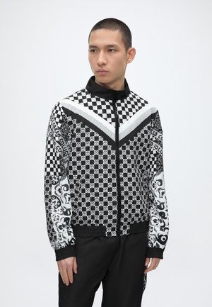 Νέος άντρας με κοντά μαλλιά, που φοράει ένα μαύρο-άσπρο καρό τζάκετ με φερμουάρ και γραφικά σχέδια και μαύρο παντελόνι, στέκεται μπροστά σε λευκό φόντο.