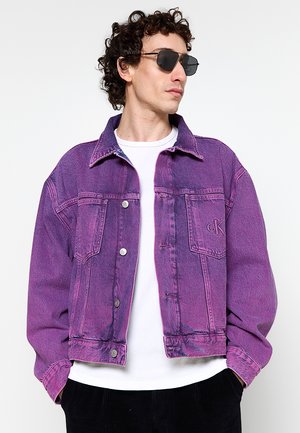 Uomo con capelli ricci che indossa occhiali da sole neri e una giacca di jeans viola sopra una camicia bianca su uno sfondo semplice.