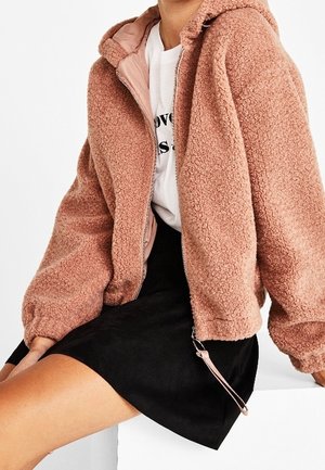 Osoba nosząca różową kurtkę teddy z polaru na białym graficznym t-shircie i czarnej spódnicy, siedząca z rękami spoczywającymi na białej powierzchni.