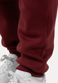 Detailní záběr na dolní část nohou v burgundských teplácích a bílých teniskách se zavázanými tkaničkami, na jednoduchém světlém pozadí.