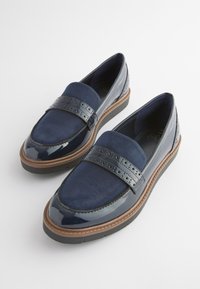Paire de mocassins bleu marine avec dessus en daim, bride en cuir perforé, bouts brillants, et semelles marron cousues sur fond blanc.