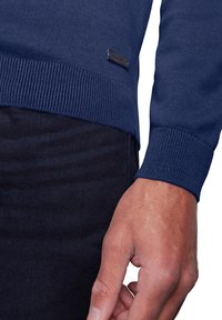 Marineblauer Strickpullover mit gerippten Bündchen und Saum. Verfügt über ein kleines schwarzes Etikett an der Seite. Kombiniert mit dunkelblauen Jeans.
